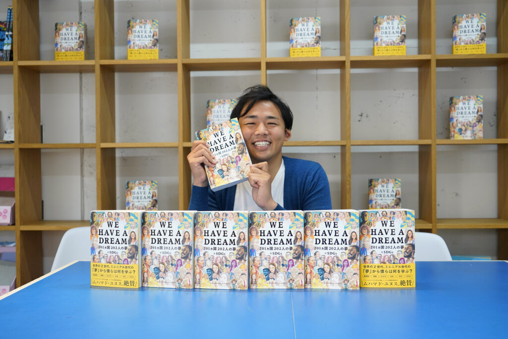 Ichikawa smiling in front of a table of copies of WE HAVE A DREAM
